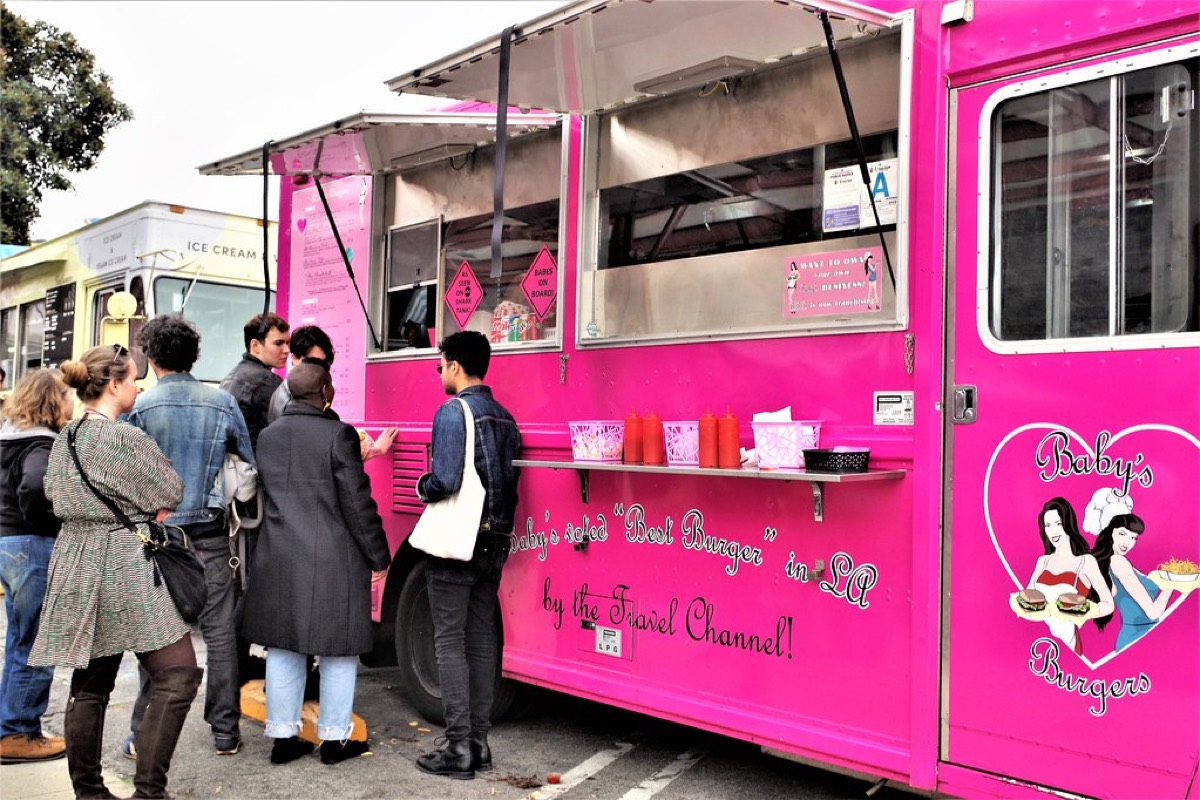 Customers lining up to order at a bright pink food truck window on a city street