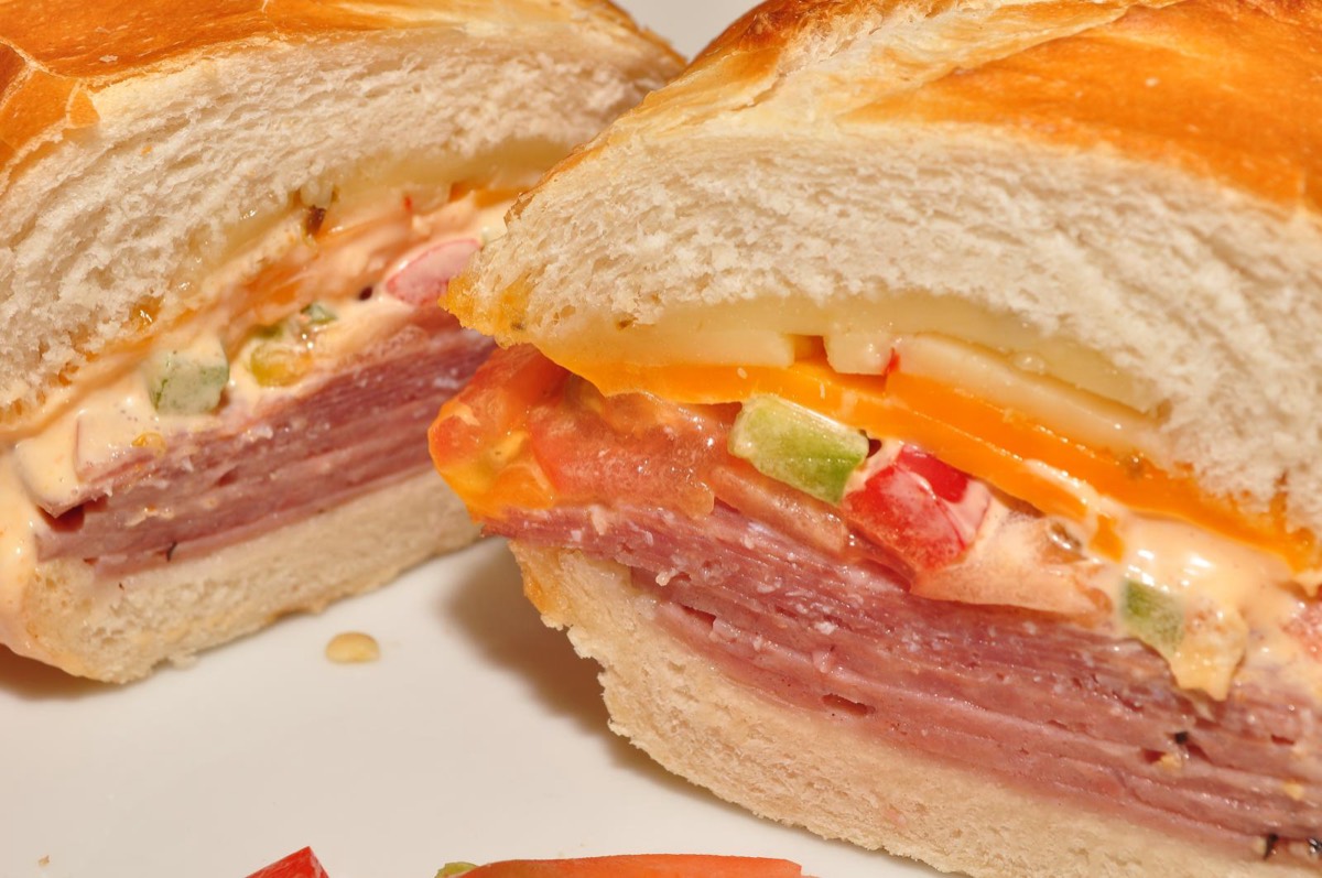 Close-up cross-section of a deli sandwich stacked with ham, salami, cheese, peppers and mayo on a fresh bread roll