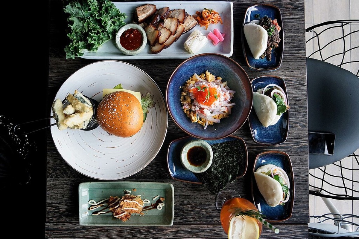 Aerial view of a restaurant table covered in many dishes, including a burger, bao buns, a rice bowl, a barbecue platter, and a small dessert, on dark wood.