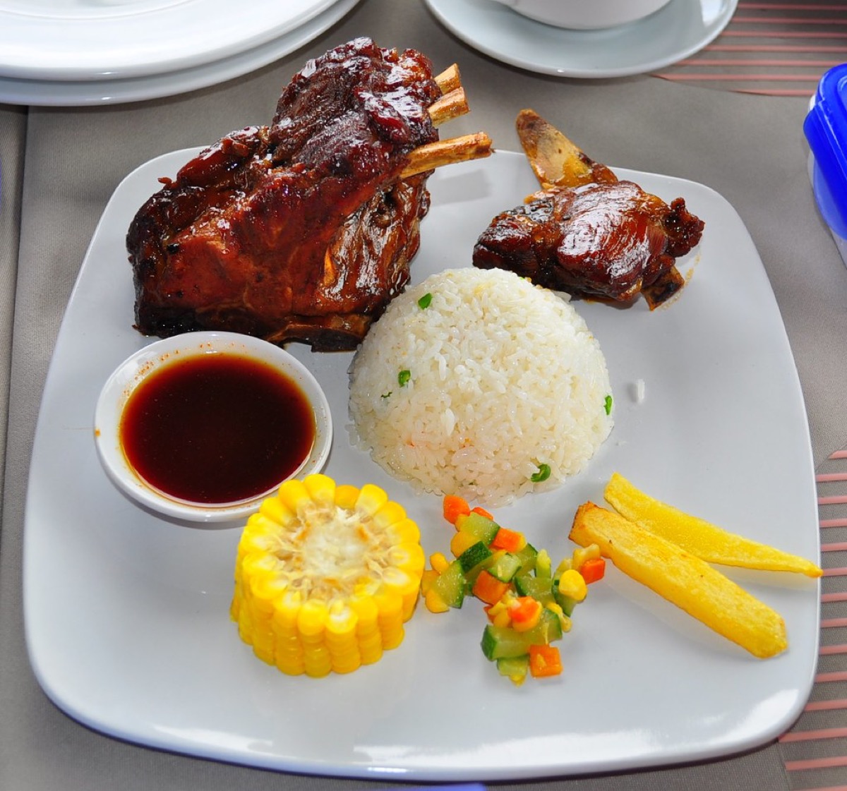 An overhead view of a restaurant plate with a rack of glazed ribs, a rice mound, corn on the cob, diced vegetables, fries and a small dipping sauce, showing a fuller order with sides added.