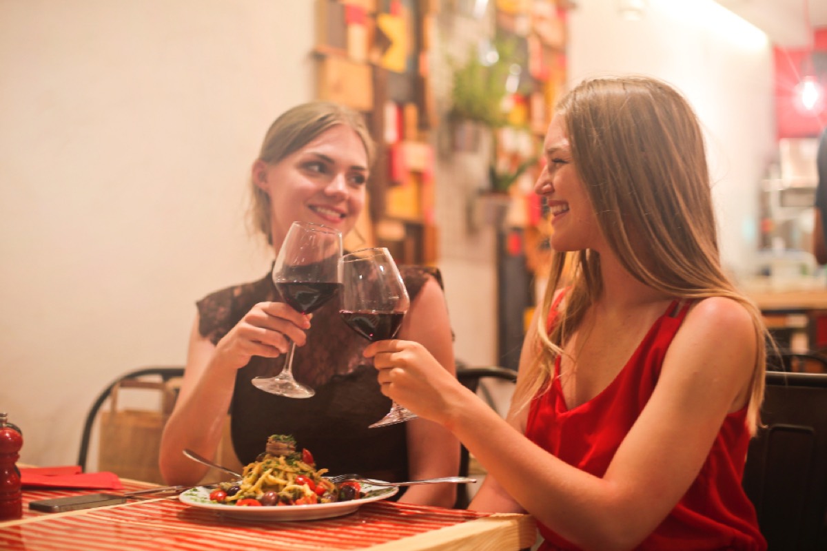 Two women in a warmly lit restaurant clinking red wine glasses together over a plate of pasta, both smiling.