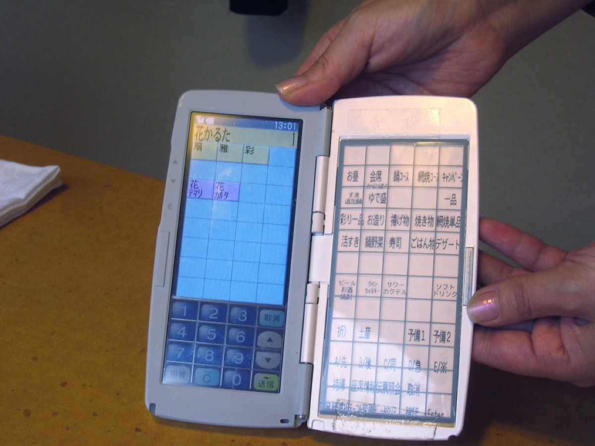 Two hands hold a folding electronic order pad with a Japanese-language menu grid used for entering food orders in a restaurant.