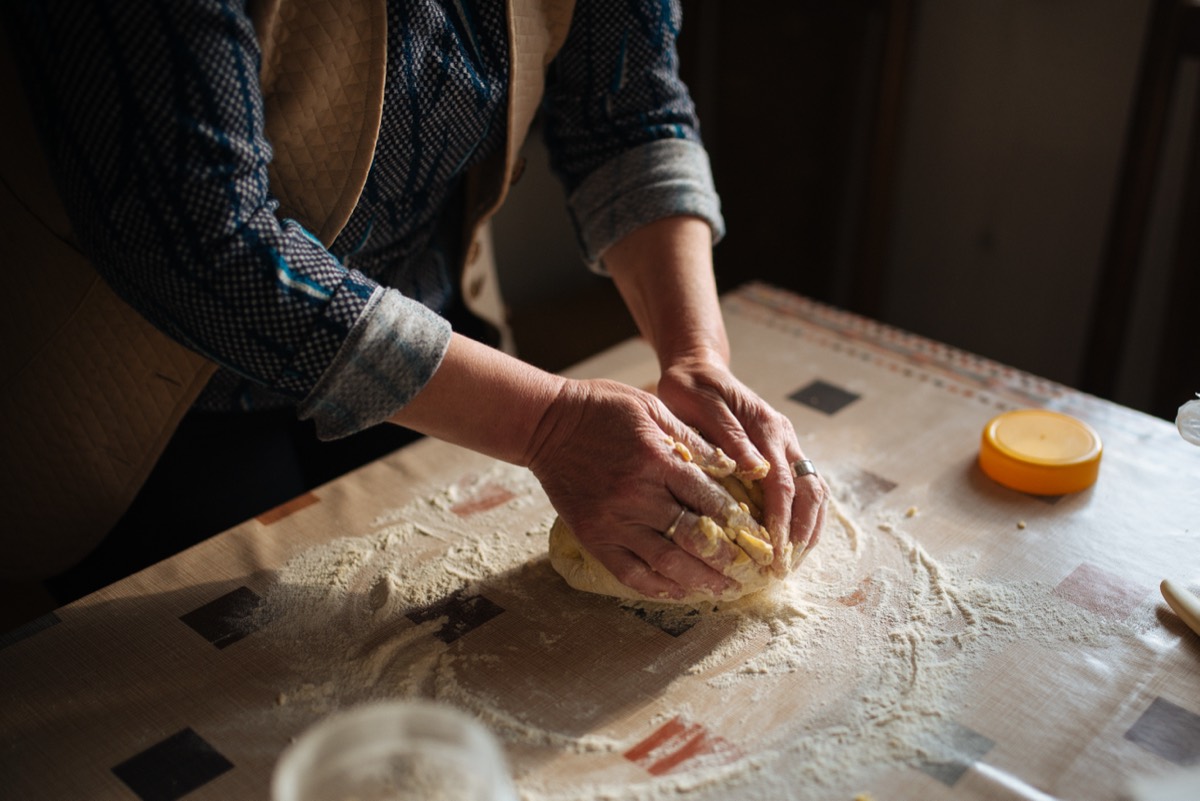 A pair of hands kneading a ball of bread dough on a floured, checkered tablecloth-covered kitchen table