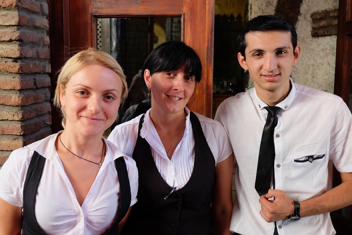 Three smiling hospitality staff, two women and one man in white shirts and black vests, standing together at a restaurant entrance.