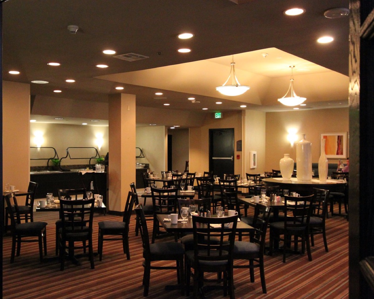 A hotel restaurant dining room with rows of black chairs and set tables, pendant lighting overhead, and a breakfast buffet station along the back wall.