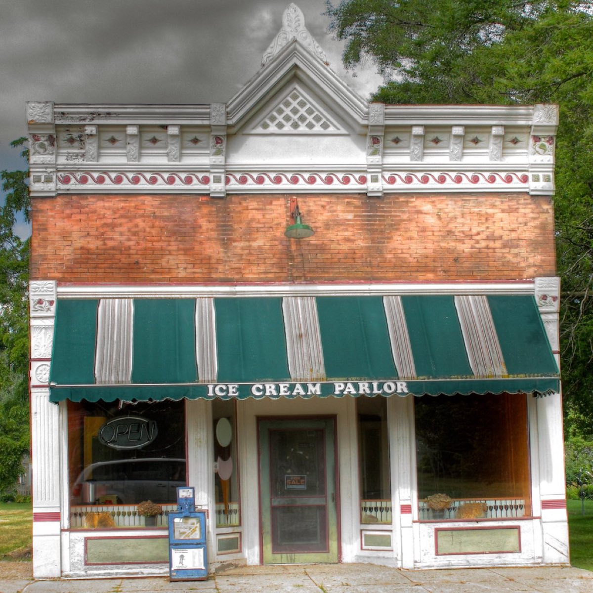 The ornate brick and white trim facade of a small town ice cream parlor, with a green striped awning reading Ice Cream Parlor above the shop windows and door.