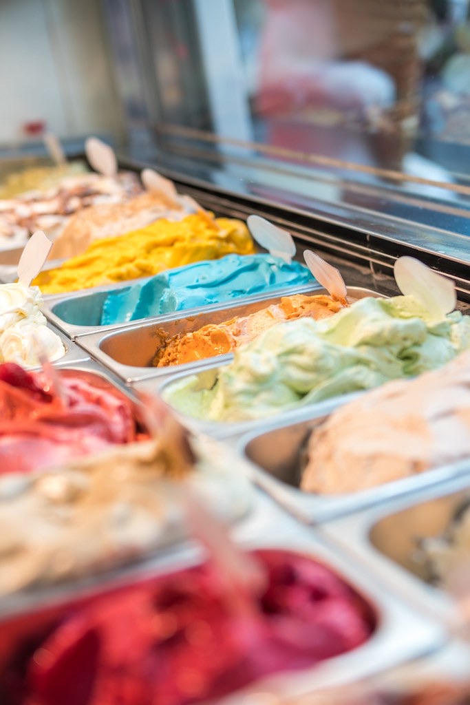 Rows of colourful gelato flavours in a metal display case with small spade scoops, shot in soft focus