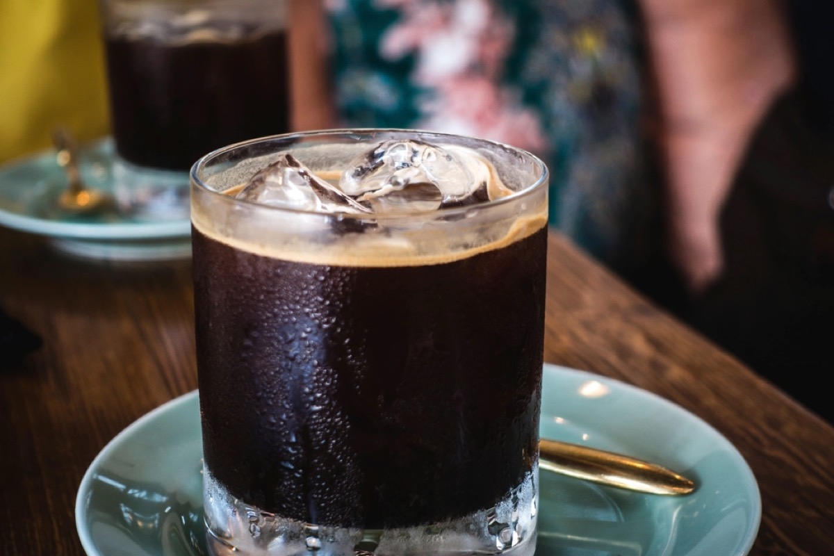 A glass of iced black coffee over ice with a crema head, on a saucer in a cafe