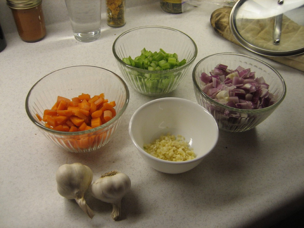 Small glass and ceramic bowls of chopped carrots, celery, red onion, and minced garlic, with two whole garlic bulbs, arranged on a kitchen counter ready for costing and cooking.