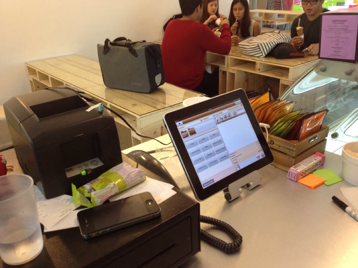 An iPad point of sale system on a stand at a small food shop counter, next to a black receipt printer and cash drawer, with customers being served in the background.