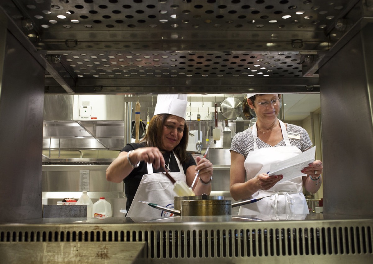 Two commercial kitchen staff in white chef coats and aprons working side by side at a stainless steel pass, one stirring a pot and one holding a docket.