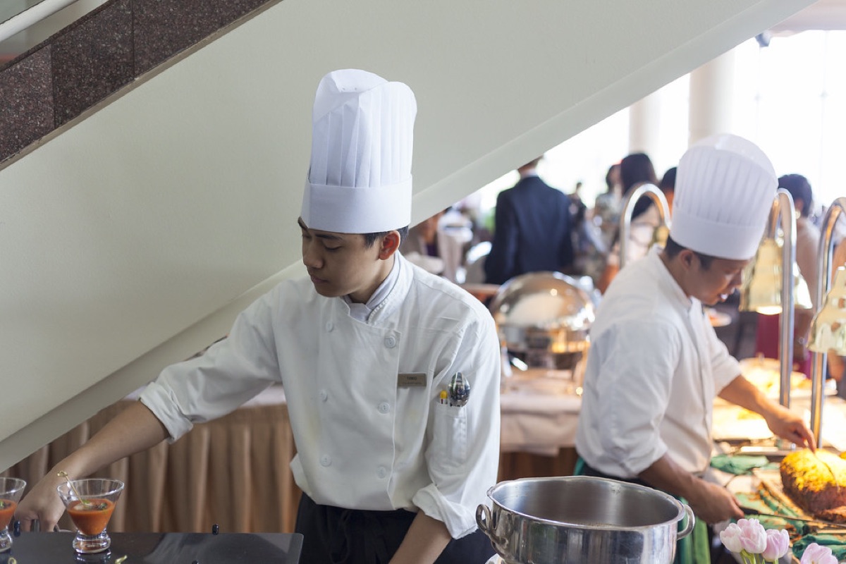 Two chefs in white uniforms and tall toques plate dishes side by side at a busy brunch buffet service.