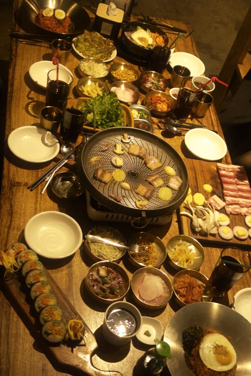 Overhead view of a Korean barbecue table with a charcoal grill cooking pork belly and mushrooms, surrounded by numerous banchan side dishes, lettuce, kimbap and drinks.
