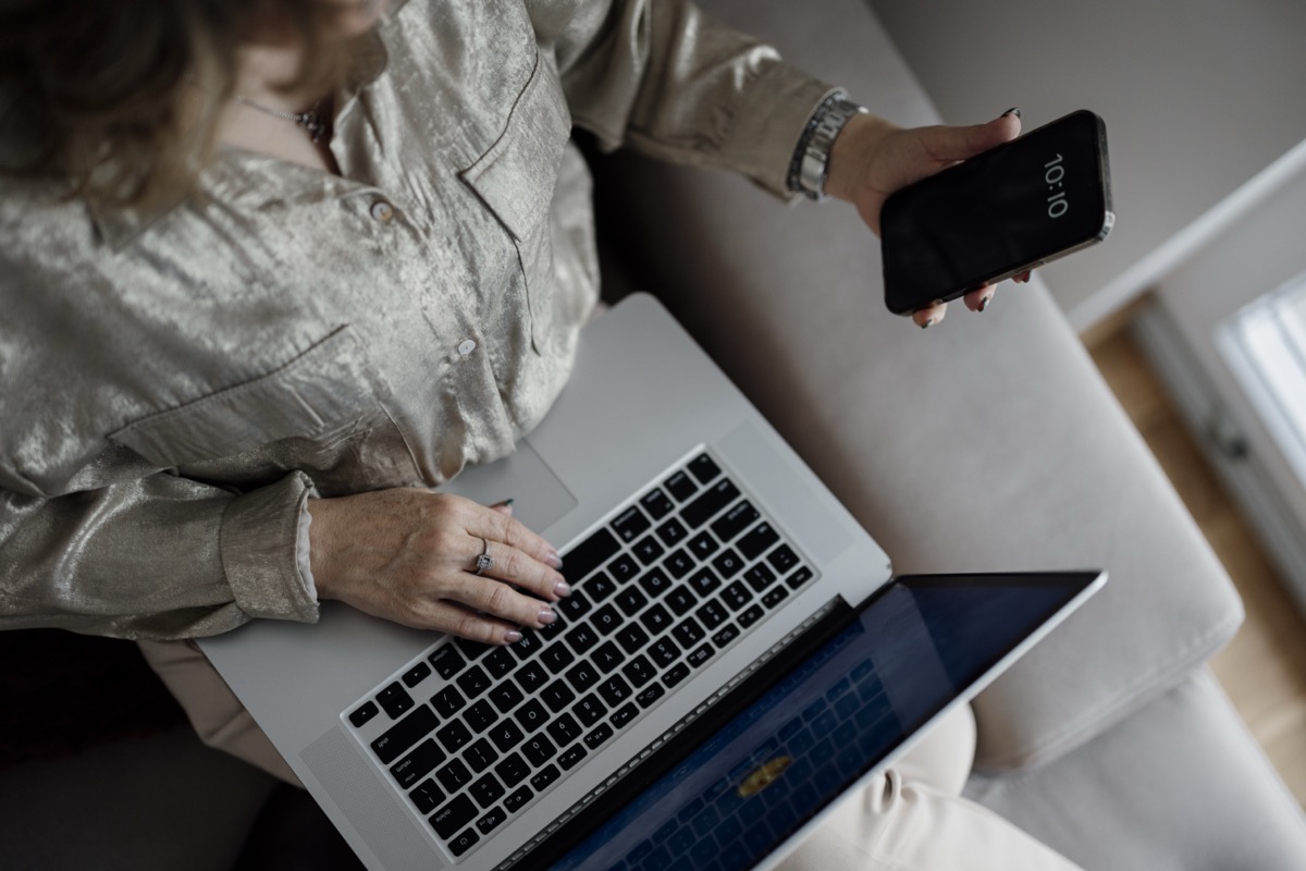 A person types on a laptop keyboard while holding a smartphone showing the time in the other hand, seated on a couch.