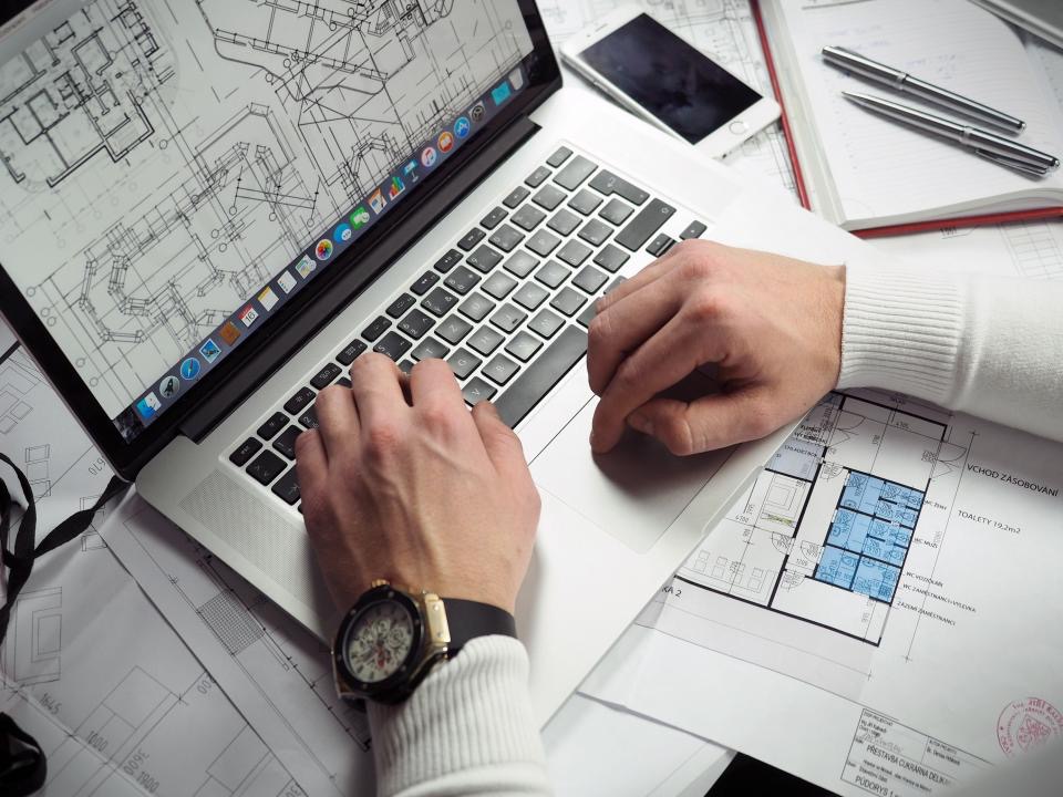 Overhead view of a person typing on a laptop showing floor plan drawings, surrounded by printed blueprints, a notebook, pens, a phone, and a wristwatch on a desk.