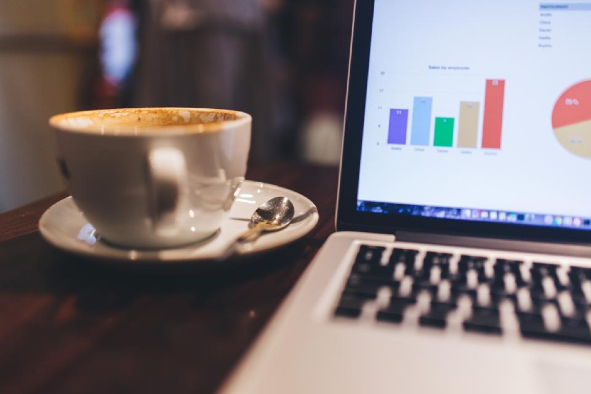 A laptop screen showing a bar chart and pie chart of sales figures, next to a coffee cup and saucer on a cafe table.