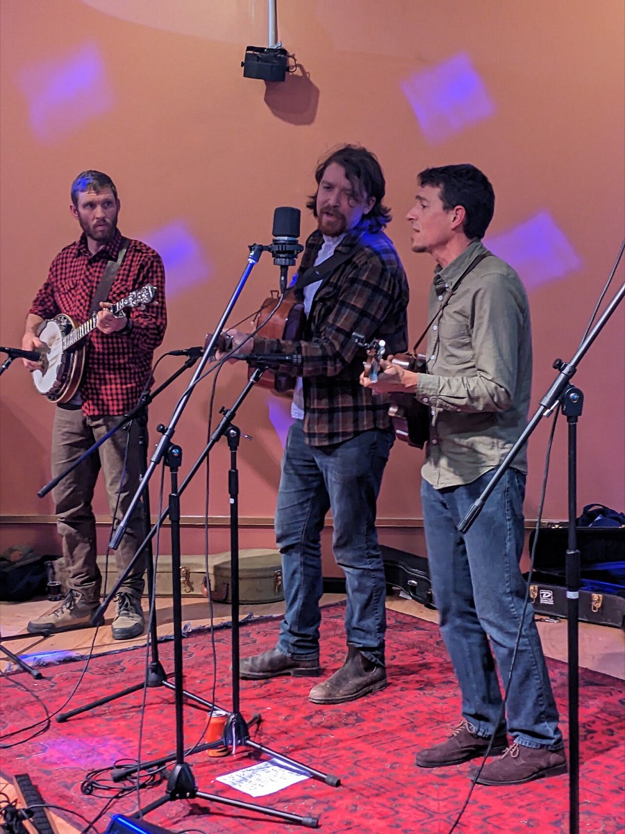 Three musicians playing banjo and guitars on a small stage with a red rug and purple stage lighting