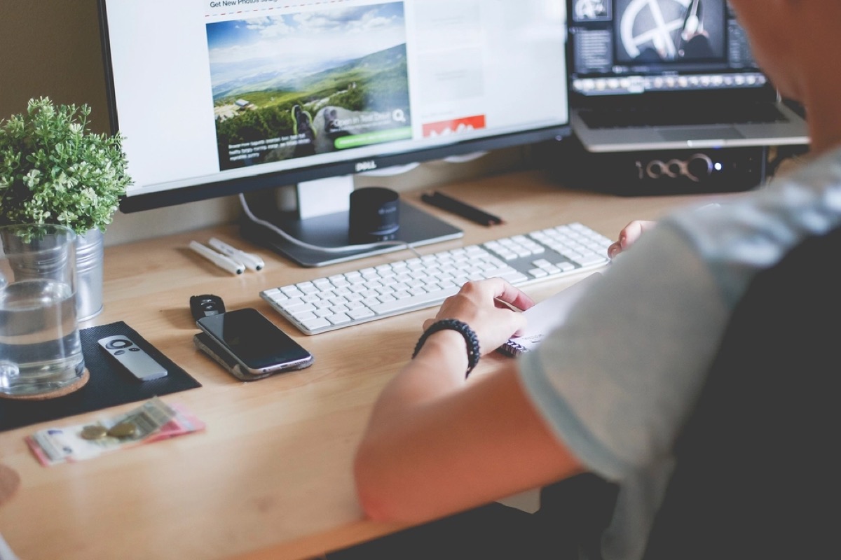 A person sits at a wooden desk typing on a keyboard, with a dual monitor setup and a laptop open showing a website layout being designed.