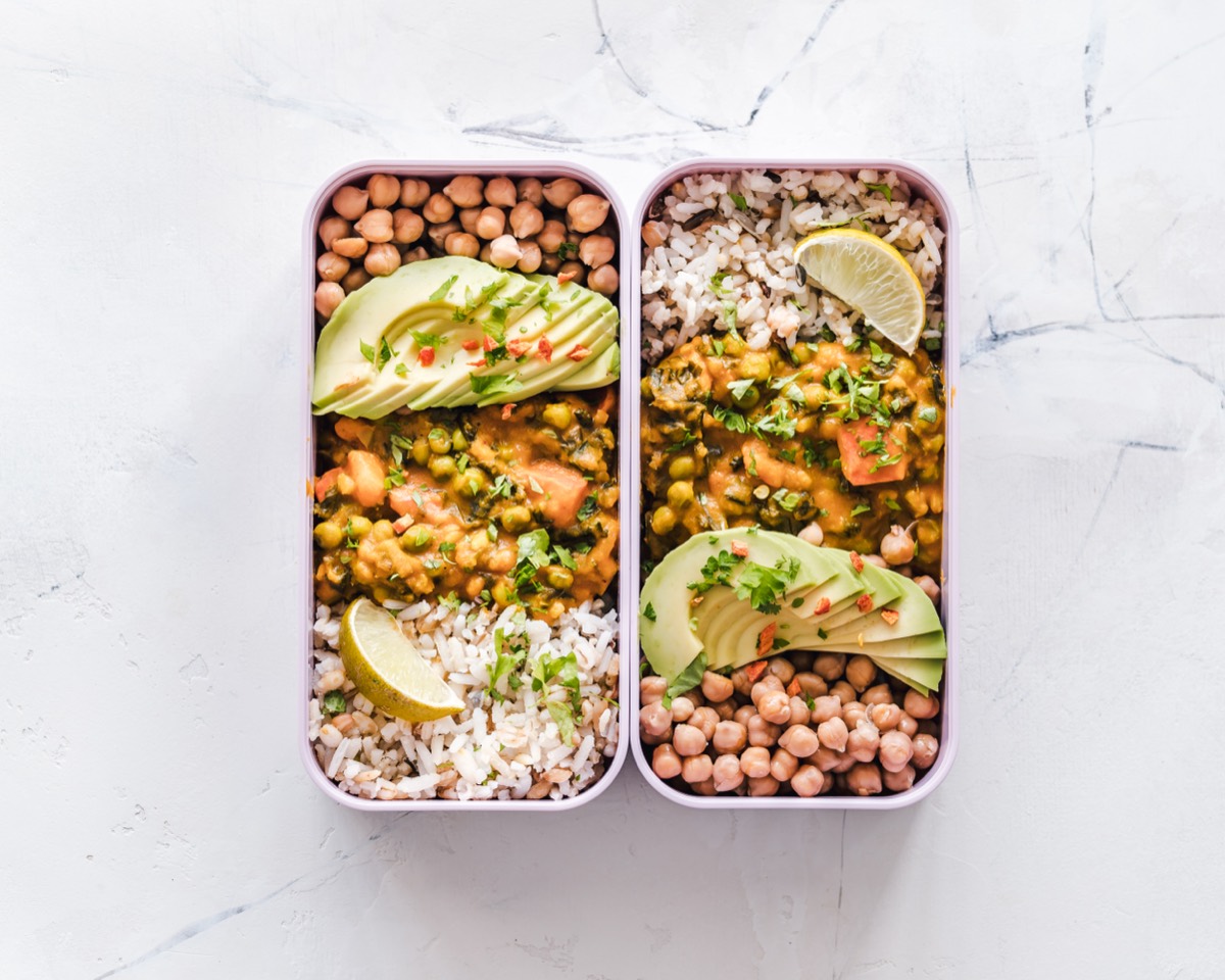 Two reusable containers filled with rice, chickpeas, avocado, and vegetable curry, viewed from above.