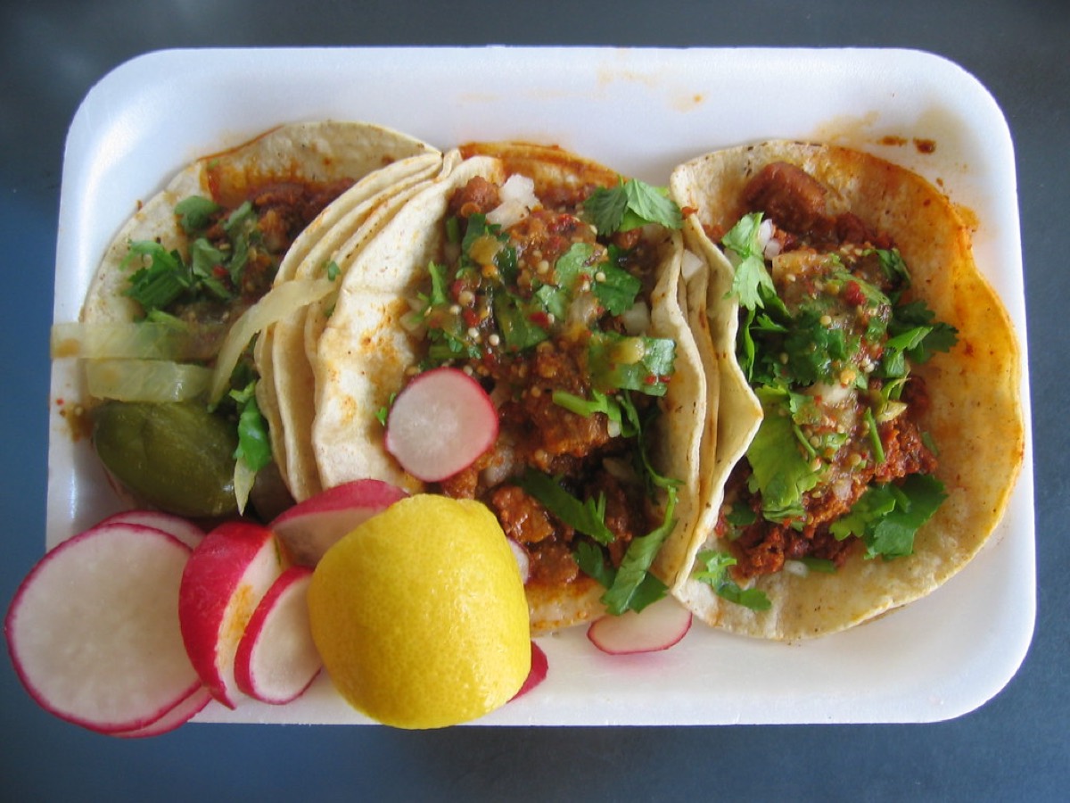 Three soft corn tortilla tacos topped with meat, onion, and cilantro, served with sliced radish, pickled jalapeno, and a whole lime.