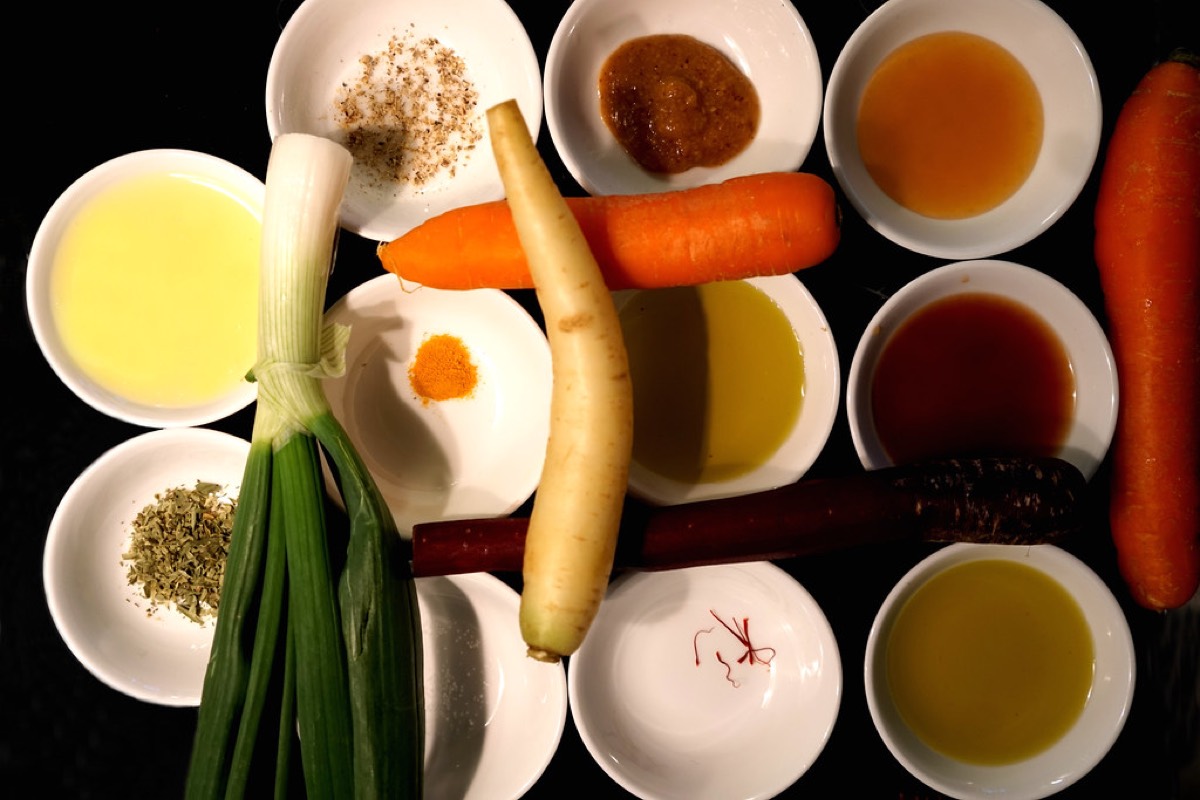 Overhead view of small white prep bowls holding chopped herbs, spices, sauces and oils arranged around fresh carrots and a spring onion on a dark surface.
