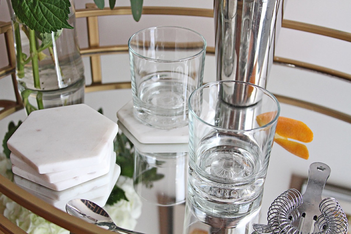 A styled bar cart topped with tumblers on marble coasters, a cocktail shaker, a strainer, fresh herbs and an orange twist