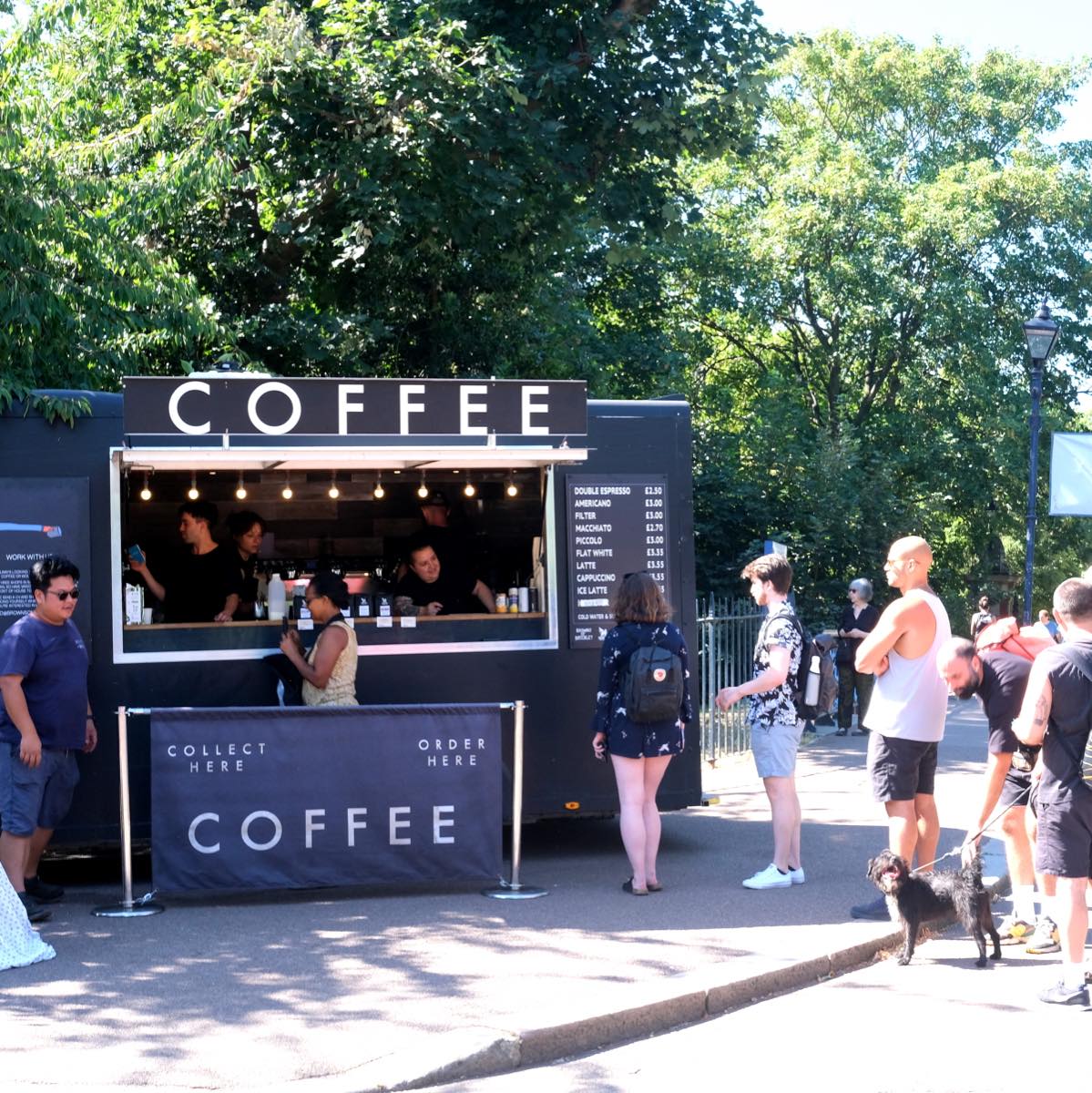 Baristas serving customers from a black mobile coffee trailer with a lit up COFFEE sign in a sunny park