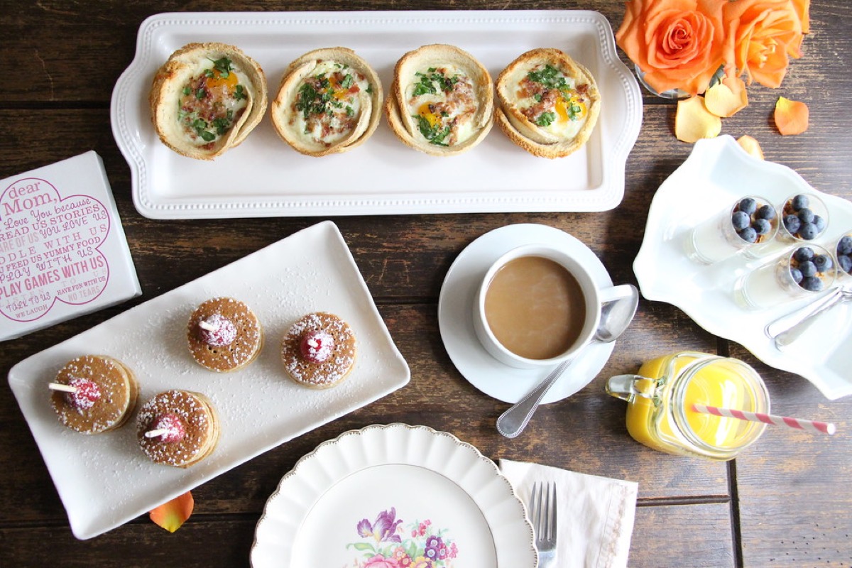 An overhead Mother's Day brunch spread with baked egg cups, mini pancakes, yogurt parfaits, coffee, orange juice and a rose