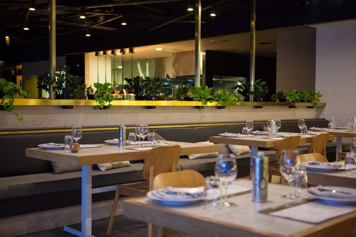 An empty modern restaurant dining room with marble topped tables set with wine glasses and cutlery, wooden chairs, and a low green plant divider under warm pendant lighting.