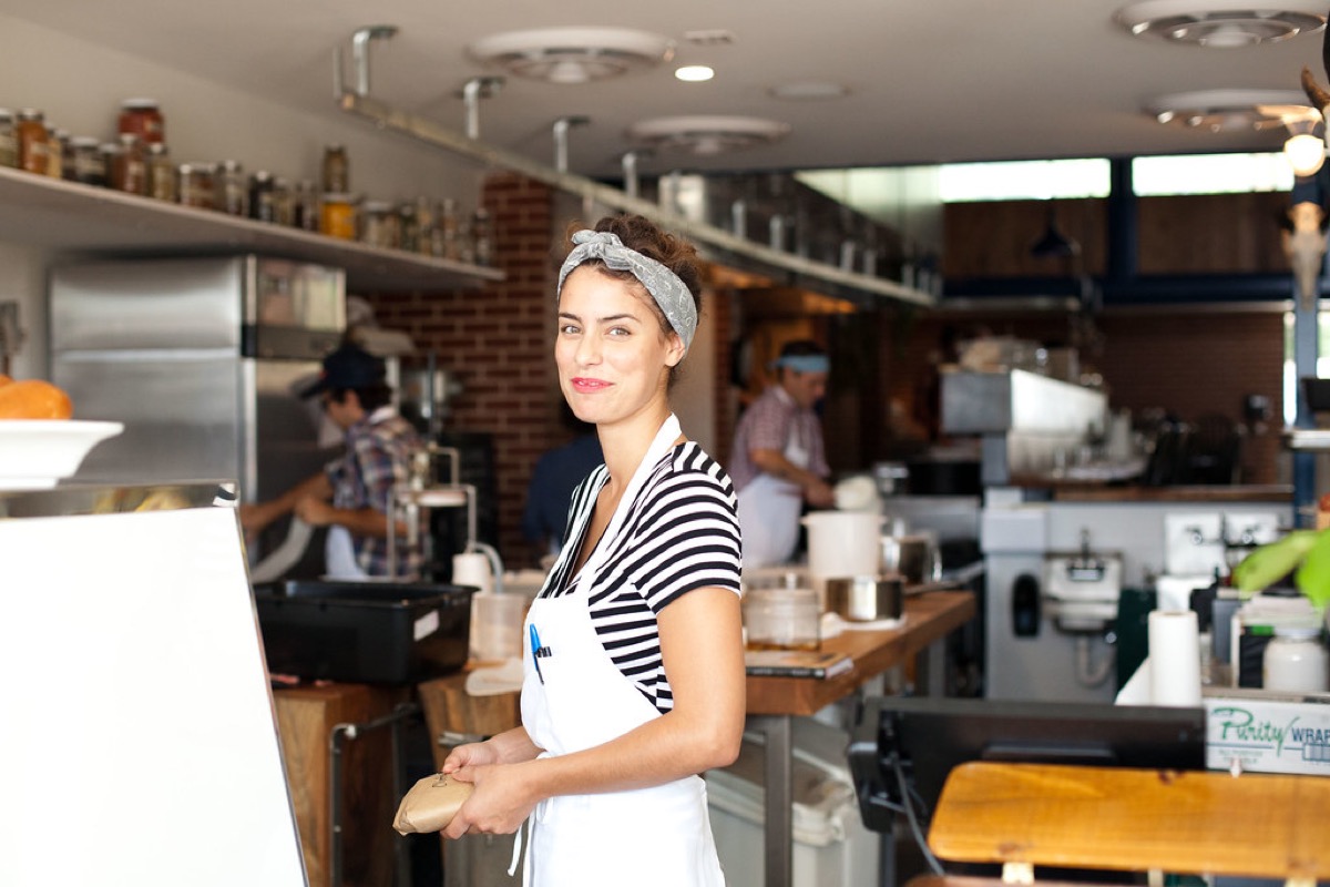 A woman in a striped shirt and white apron stands smiling at the counter of a newly opened food shop, with staff working behind her.