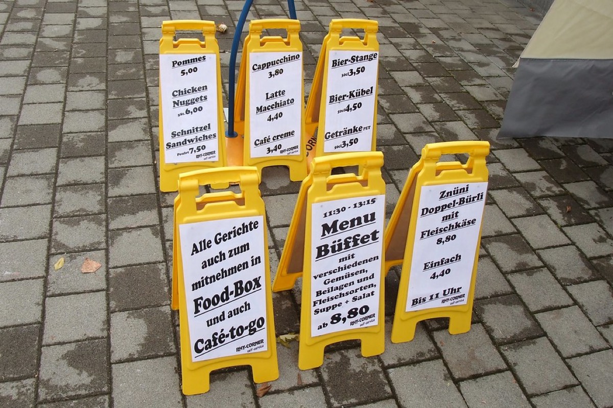 Six yellow A-frame sandwich board signs on a paved street, each listing food and drink items with clearly printed prices such as Cappuccino 3.80 and Menu Buffet 8.80.