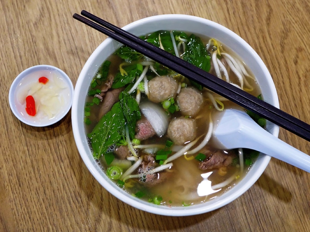 An overhead bowl of beef pho with noodles, meatballs, bean sprouts and herbs, chopsticks resting on top