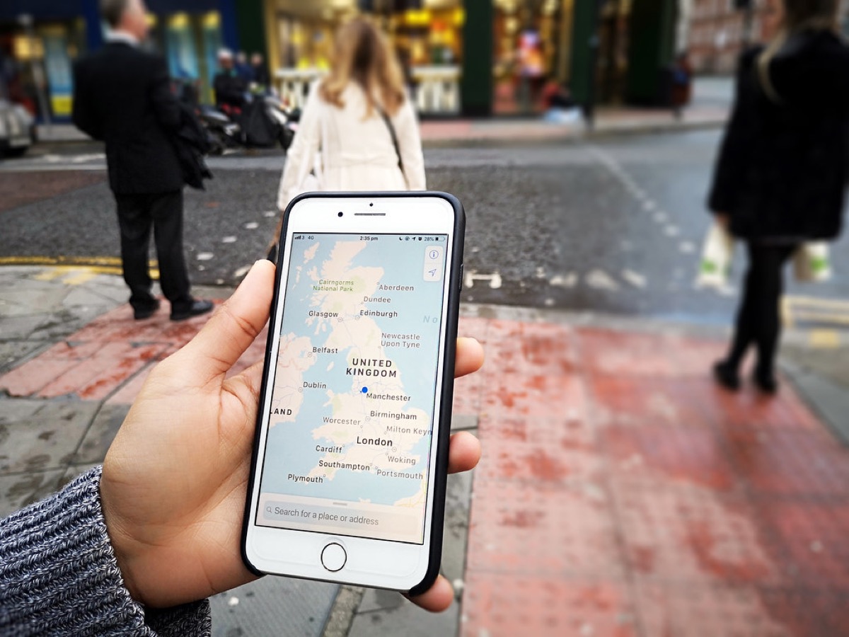 A hand holds up an iPhone showing a map app with a search bar reading Search for a place or address, on a city street with pedestrians in the background.