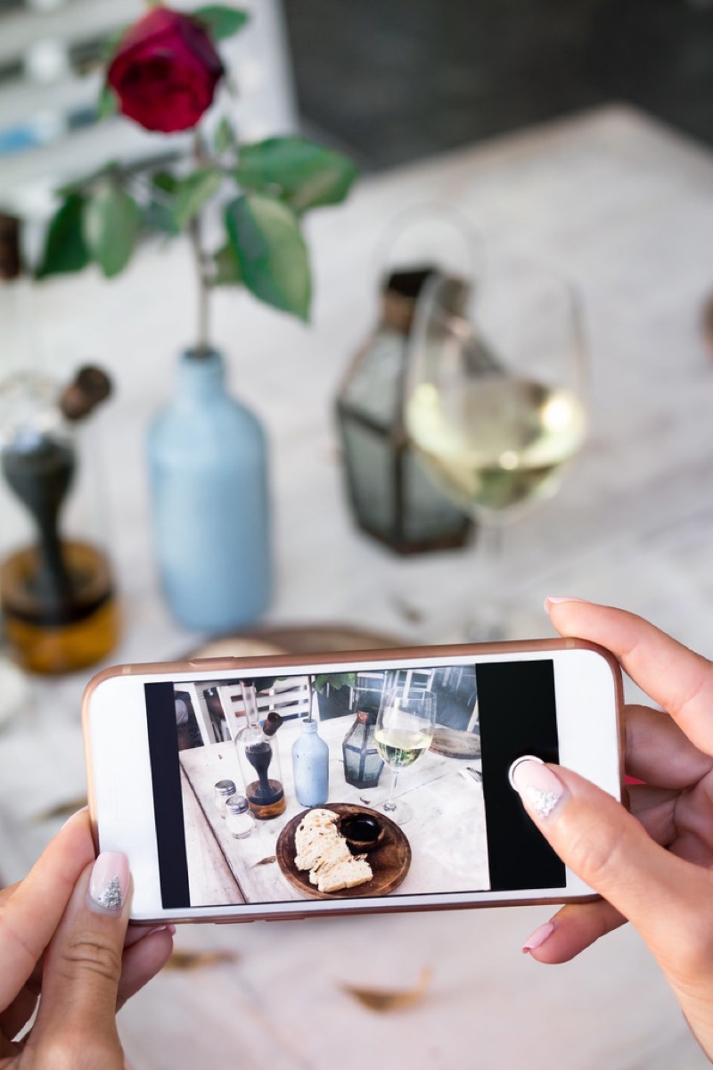 Hands holding a smartphone photographing a plate of bread and a glass of wine on a restaurant table