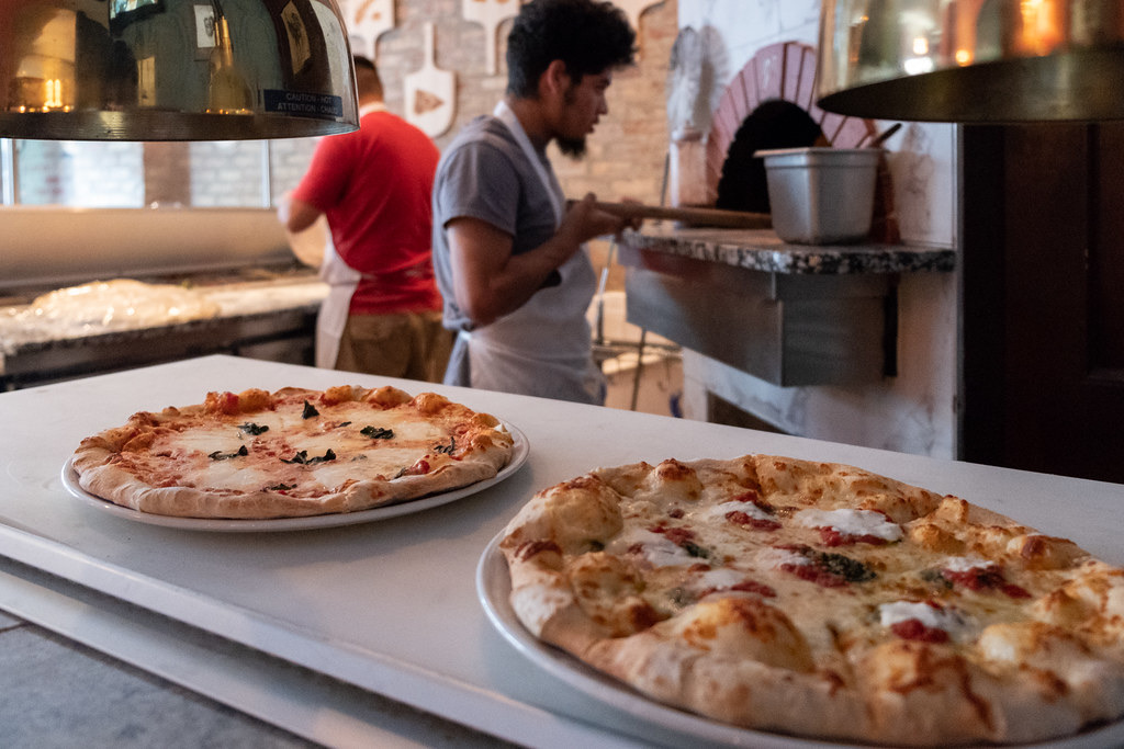 Two freshly baked pizzas on plates in the foreground while a pizzaiolo works at a wood-fired oven in the background