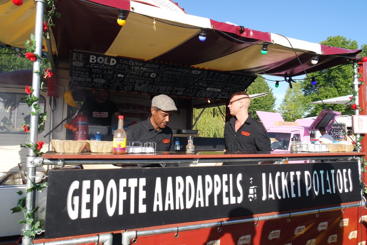 Two staff members work behind the counter of a decorated food truck at an outdoor festival under a sunny sky.