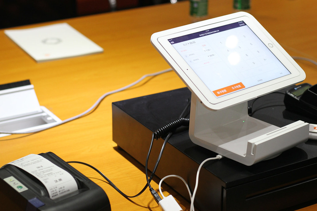 A tablet based point of sale terminal on a stand next to a receipt printer and cash drawer on a wooden counter, with a printed receipt visible.