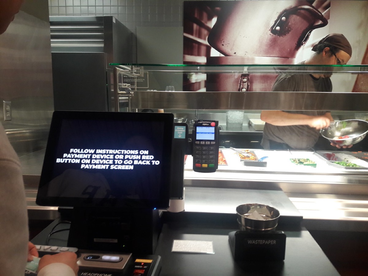 A point of sale screen and card payment terminal on a food counter, with a staff member serving behind the servery glass