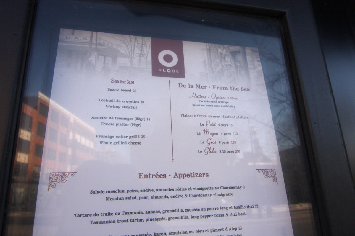 A printed restaurant menu with sections for snacks, seafood, and appetizers, including item names and prices, displayed behind glass at a restaurant entrance.