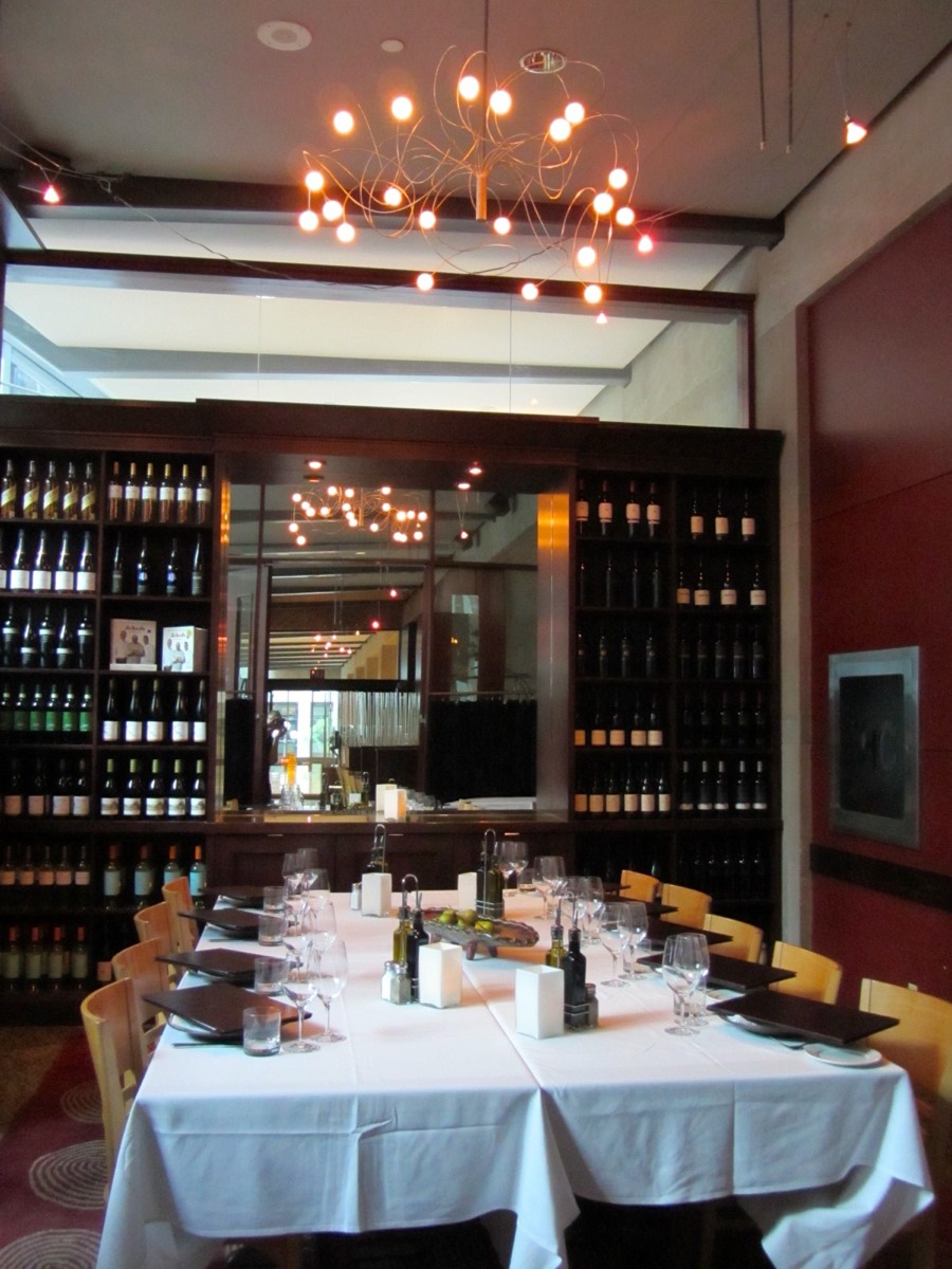 A long table set with white linen, wine glasses, and place settings inside a restaurant private dining room lined with a wine wall.