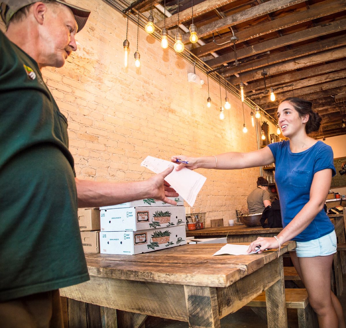A restaurant staff member in a blue shirt signs a delivery invoice handed over by a produce supplier, with boxes of fresh vegetables stacked on a wooden table inside the restaurant.