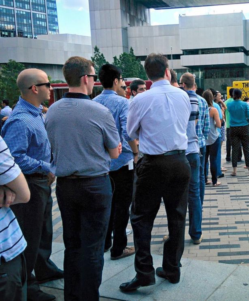 A long line of office workers stands queued outdoors, seen from behind, waiting to reach a counter to place their order.