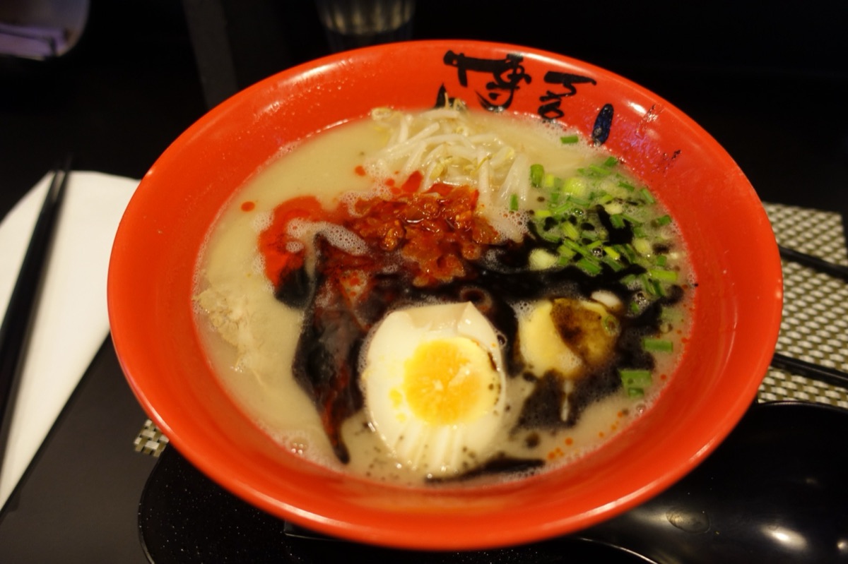Overhead view of a red bowl of tonkotsu ramen topped with a soft-boiled egg, bean sprouts, chilli, scallions and black garlic oil