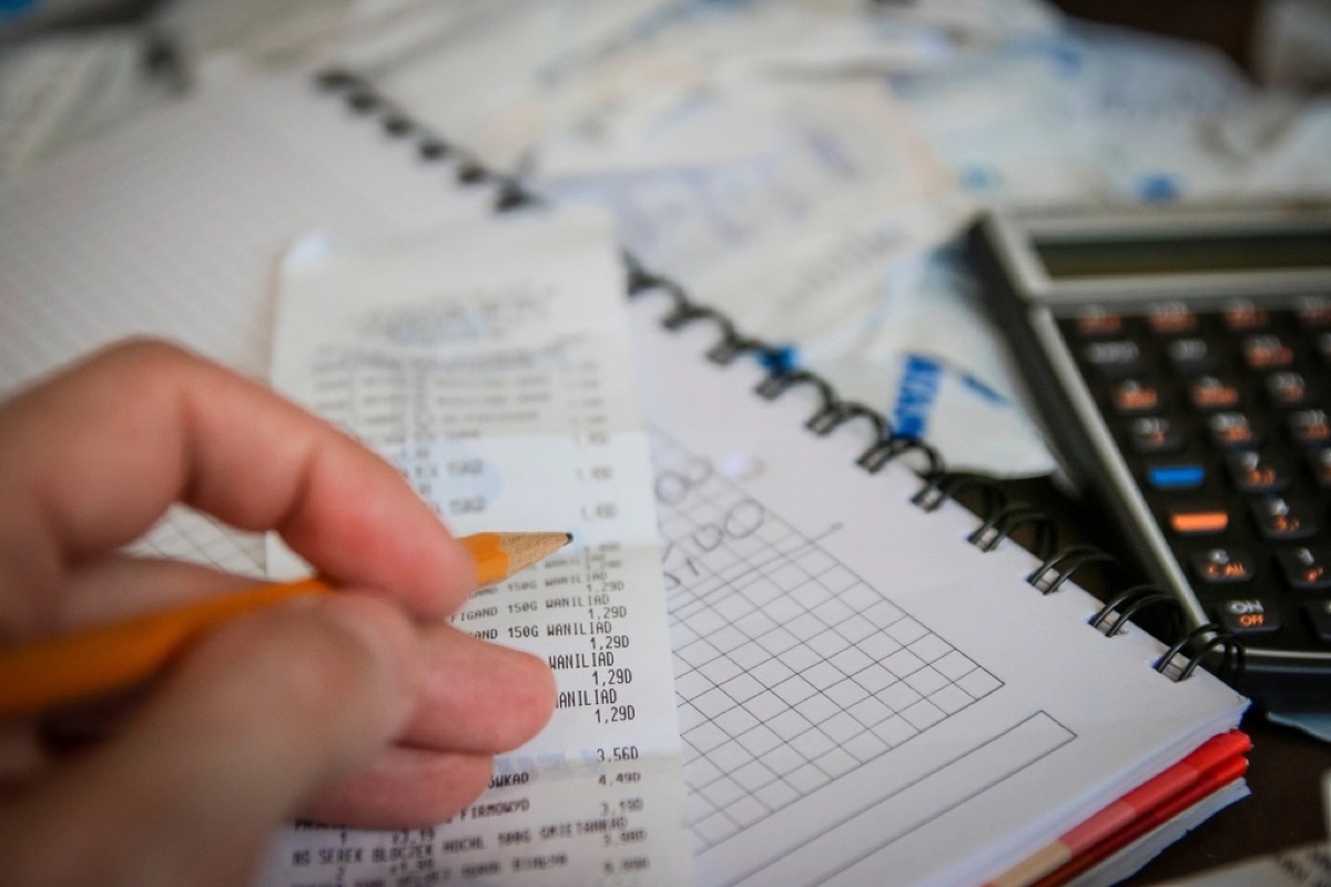A person's hand holds a pencil while checking a printed paper receipt against a spiral bound ledger notebook ruled with a grid, with a calculator visible in the background.