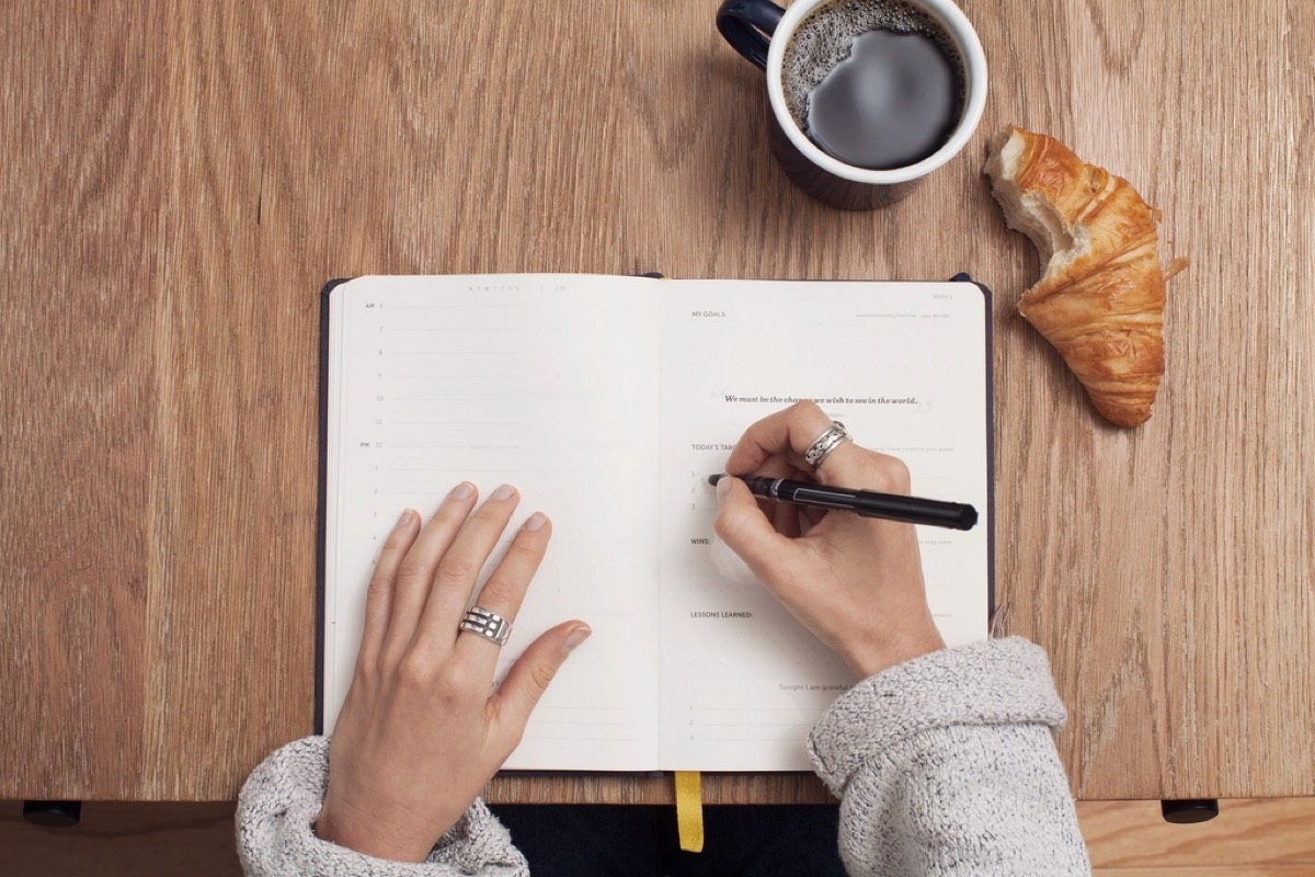 Overhead view of two hands writing in an open time-slotted diary planner, next to a mug of coffee and a partly eaten croissant on a wooden table.