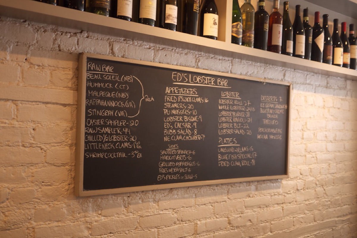 A hand-lettered chalkboard menu on a whitewashed brick wall listing raw bar, appetizers, lobster dishes and desserts with prices, below a shelf of wine bottles.