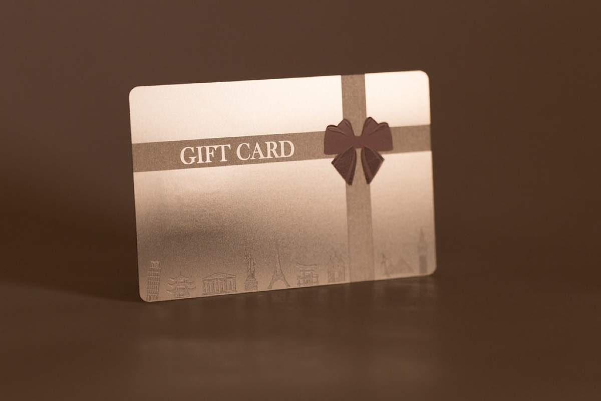 A metallic rose gold gift card standing upright on a dark surface, printed with a ribbon bow design and the words GIFT CARD