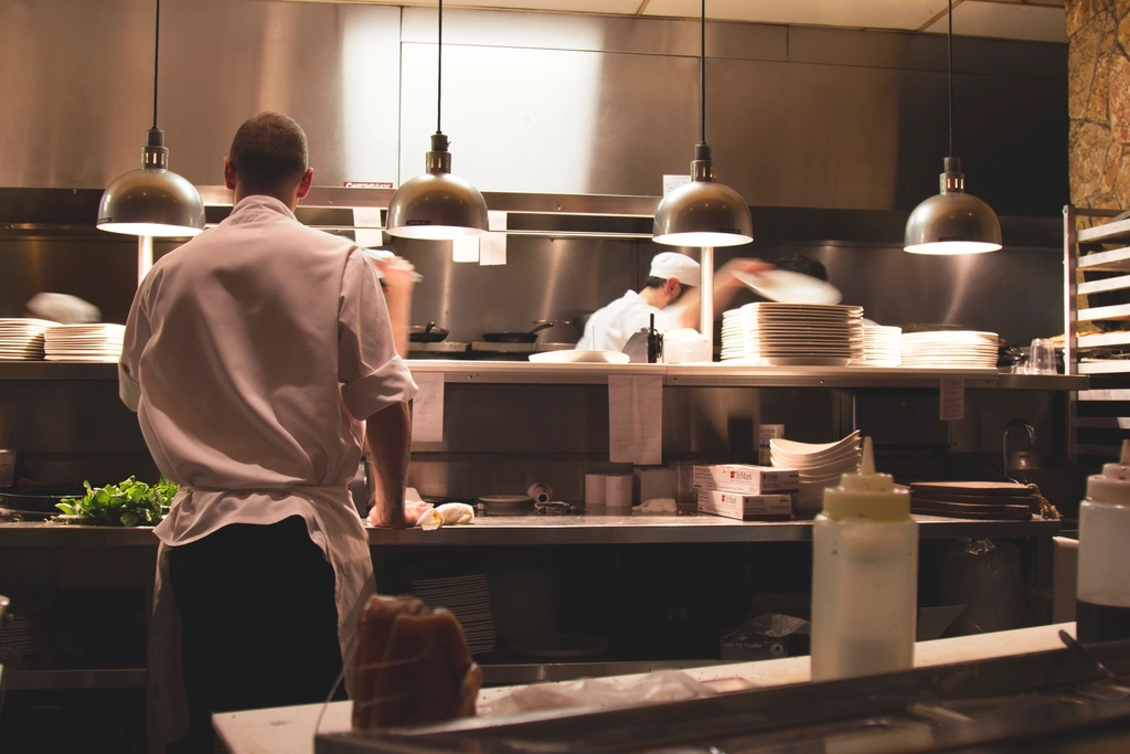 A chef stands at a restaurant kitchen pass under warm heat lamps, with stacks of white plates and cooks working the line behind
