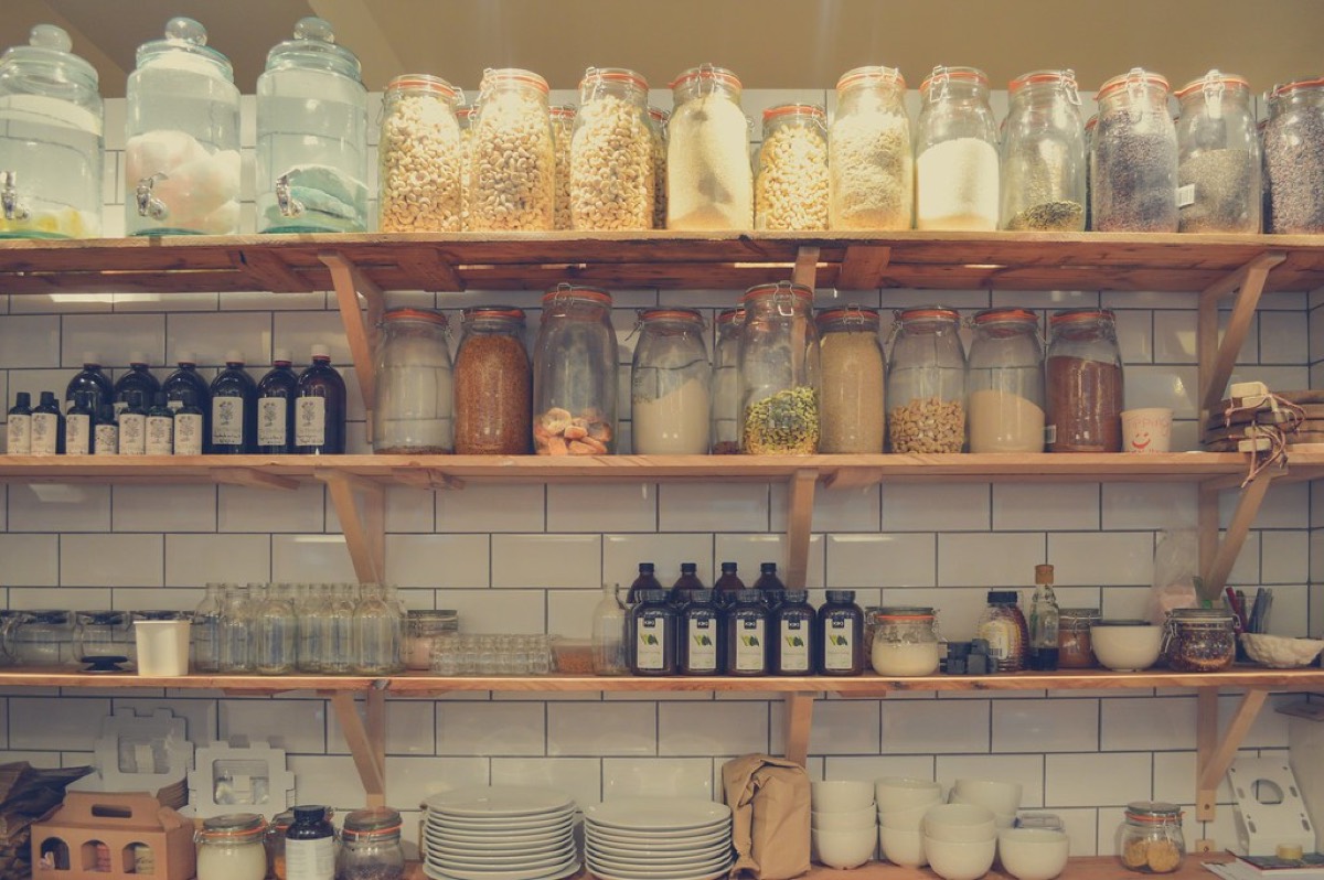 Glass jars of grains, pasta, and dried goods neatly organised on wooden shelves against white subway tile