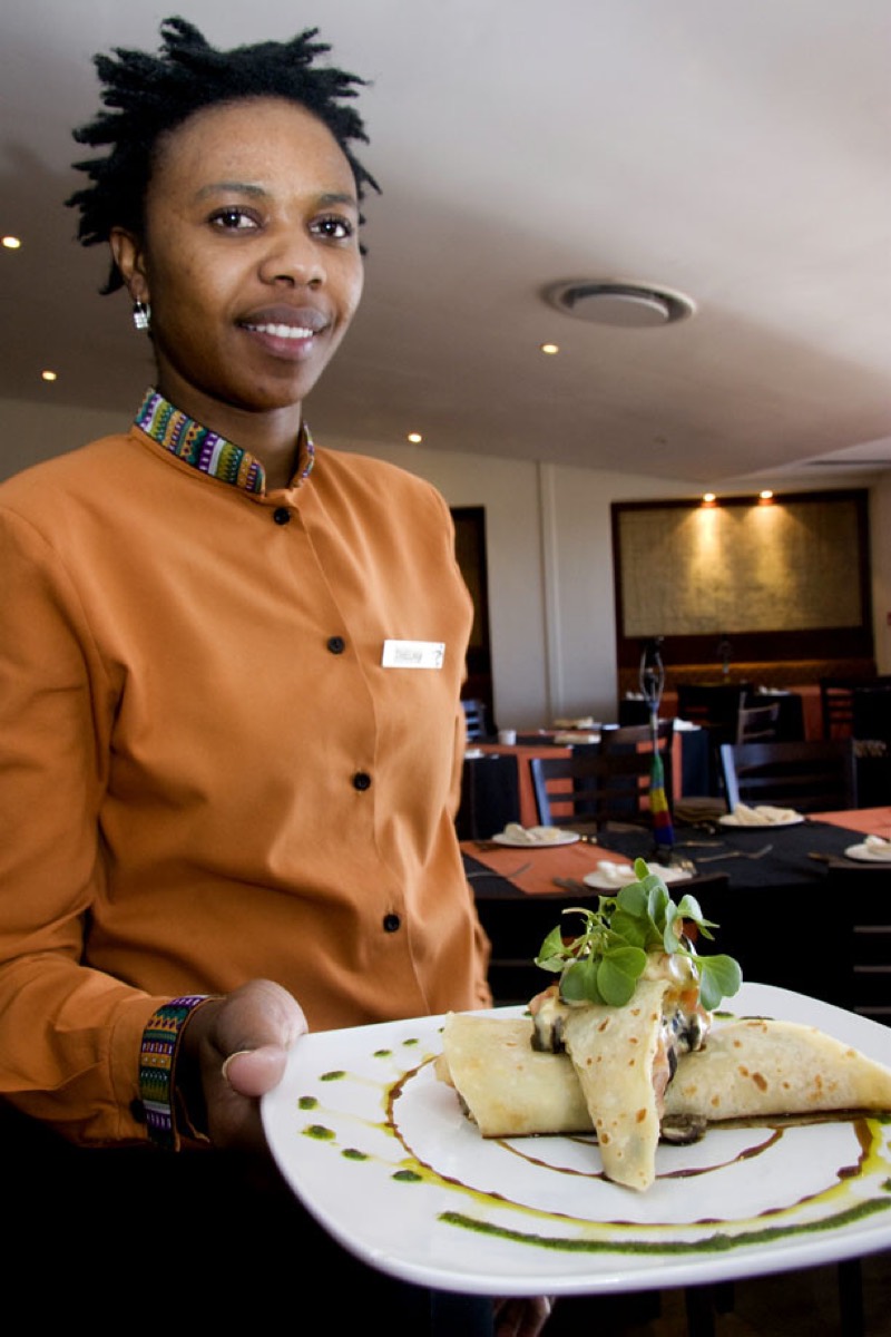 A smiling restaurant server in an orange uniform holding a white plate with a garnished crepe dish, dining room tables visible in the background